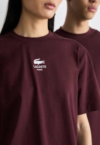 Burgundské bavlněné tričko s bílým logem Lacoste a textem "PARIS". Klasický střih s krátkými rukávy a designem s kulatým výstřihem.