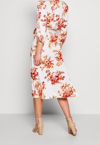 Robe florale blanche avec motifs de roses rouges et jaunes, manches trois-quarts, taille ceinturée, et jupe fluide. Portée avec des talons beiges.
