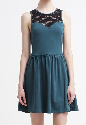 Robe sans manches couleur sarcelle avec jupe froncée et panneau de dentelle noire traversant le haut de la poitrine et les épaules, portée par une personne debout.