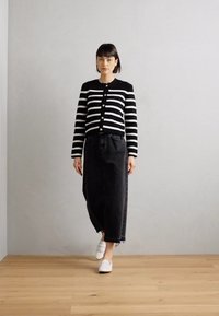 Black and white striped cardigan with round neckline and buttons, paired with high-waisted black skirt and white slip-on shoes. Wooden floor background.
