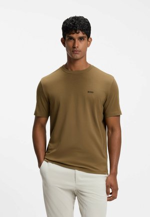 Ung mand med kort krøllet hår iført en brun BOSS t-shirt og lyse bukser, står med den ene hånd i lommen.