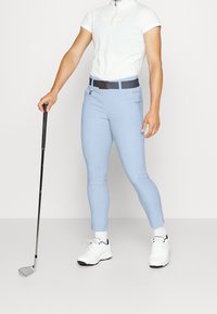 Helle blaue Golfhosen mit schlankem Schnitt, Gürtelschlaufen und Vordertaschen; kombiniert mit einem weißen Zip-up-Polo und weißen Sportschuhen.