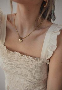 Collier en chaîne dorée avec un pendentif en forme de cœur, porté sur un haut écru à smocks avec des bords froncés et des bretelles.