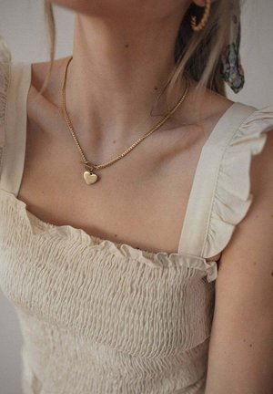 Een gouden ketting met een harthanger, gedragen over een lichtbeige, gesmokt top met ruches en banden.