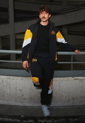 Marineblauw en geel trainingspak met kleurvlakken; jas heeft witte strepen, elastische manchetten en logo. Model staat tegen een betonnen barrierleuning.