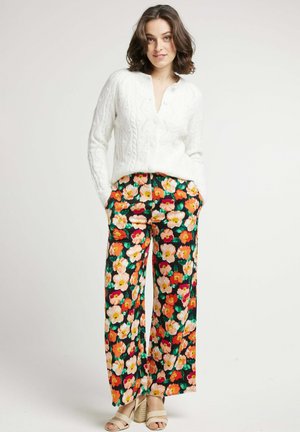 Cardigan blanc en tricot torsadé avec col rond et fermeture par boutons, associé à un pantalon large à fleurs avec des fleurs orange et rose sur fond noir.