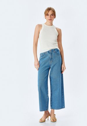 Hvid ribbet ærmeløs top med høj hals, parret med højtaljede, bredbenede blå denim jeans og gule strop hæle.