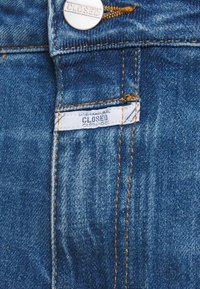 Džíny z denimu s tmavě modrým odstínem, zdobené oranžovým šitím, stříbrným knoflíkem a bílou štítkem přišitým do kapsy.