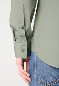 Personne portant une chemise à manches longues vert clair avec des poignets boutonnés et un jean en denim bleu, main détendue sur le côté.