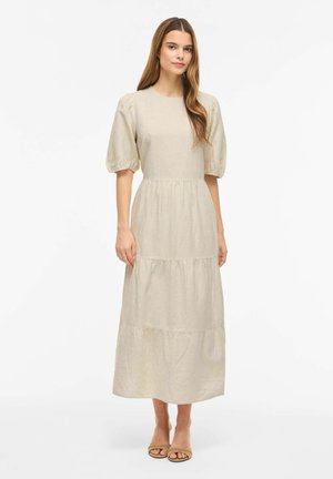 Junge Frau trägt ein beiges, gestuftes Midikleid mit Puffärmeln und offenen Sandalen, steht vor einem weißen Hintergrund.