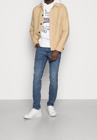 Beige linne skjortjacka över en vit hoodies med blå tryck, matchad med blå jeans och vita sneakers på ett grått golv.