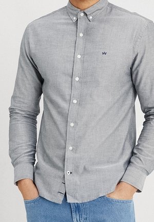 Mann trägt hellgraues Hemd mit kleinen dunkelblauen bestickten Logo auf der Brust, kombiniert mit blauer Jeans, die Hände in den Taschen.