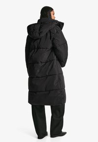 Svart vadderad jacka med huva, med ett quiltat mönster och en midjelång design. Bärs över mörka byxor och har en slät textur.