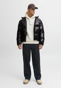 Uomo che indossa una giacca a vento nera, una felpa bianca in pile, pantaloni neri, stivali beige e un cappello scuro, mentre si trova davanti a uno sfondo chiaro e semplice.