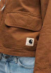 Καφέ τσέπη μπουφάν Carhartt με καπάκι και λογότυπο ακριβώς πάνω από τη μέση μπλε τζιν και θηλιά ζώνης.