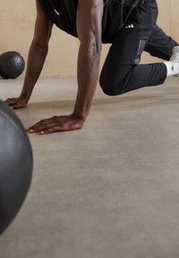 Idrottare i svart träningsklädsel, placerad på händer och knän på en grå yta, med en svart medicinboll i närheten.