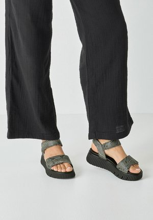 Grønne sandaler med tekstur og brede remme, platåsål og kontrasterende sort gummibund, kombineret med flagrende sorte bukser.