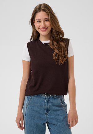 Jeune femme aux longs cheveux bruns sourit, portant un haut noir sans manches par-dessus un T-shirt blanc et un jean bleu avec une ceinture noire.