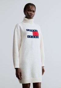 Fehér kötött ruha magas nyakrésszel. Bő ujjakkal és bordázott szegéllyel rendelkezik. Egy tengerészkék, piros és fehér színű logófoltot mutat.