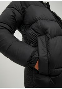 Osoba w czarnej pikowanej kurtce puchowej z ręką w bocznej kieszeni, widoczna od klatki piersiowej do nadgarstka.