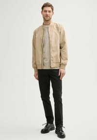 Cazadora bomber beige con cremallera frontal y puños acanalados, llevada sobre una camiseta gris claro. Combinada con pantalones negros y zapatos negros de cordones.
