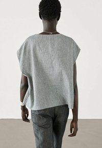 Gray, textured top with short sleeves and a loose fit, featuring a back closure. Paired with dark, patterned trousers.