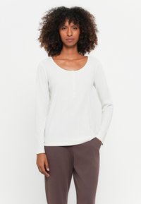 Vrouw met krullend haar, gekleed in een witte lange mouw henley-shirt en bruine broek, met één hand in de zak, staand tegen een effen witte achtergrond.