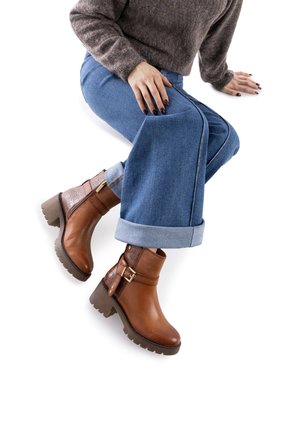 Botas de cuero marrón hasta el tobillo con un acento texturizado, un zipper lateral y una suela gruesa con tacos. Llevadas con vaqueros azules doblados.