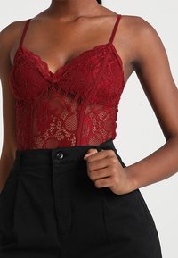 Viininpunainen pitsinen vartalopuku, jossa on herkät kukkakuviot ja säädettävät ohuet olkaimet, yhdistettynä korkeavyötäröisiin mustiin housuihin.