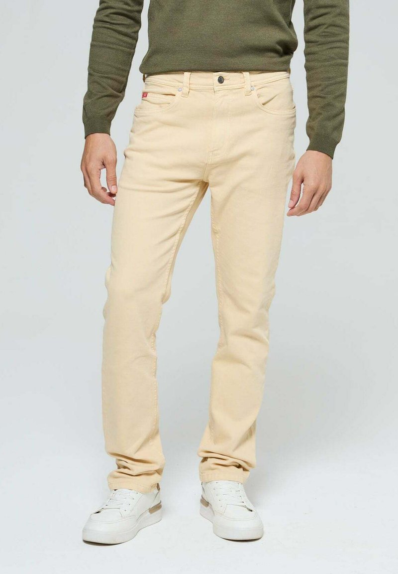 Pantalons en coton beige à coupe ajustée, avec cinq poches, des passants de ceinture et une fermeture à bouton. Portés avec des baskets blanches.