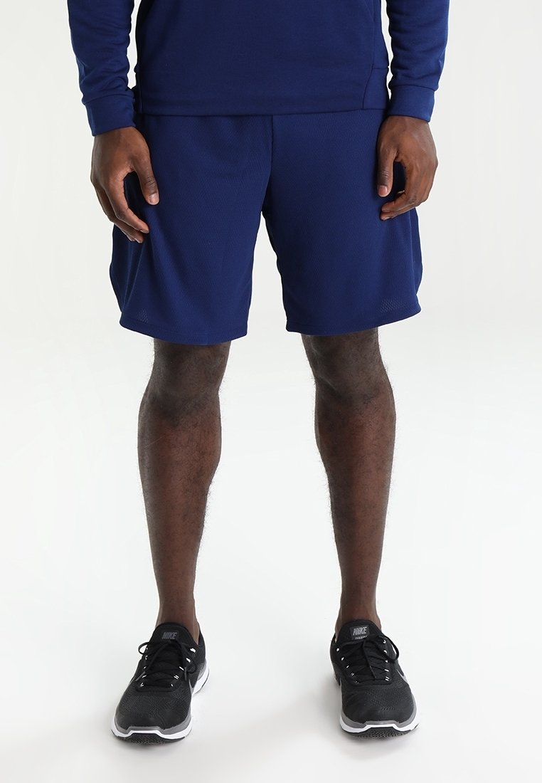 Homme portant un short de sport bleu marine, un haut à manches longues assorti et des chaussures de course Nike noires, debout sur un fond blanc.