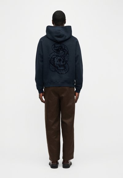 Homme debout de dos, portant un sweat à capuche sombre avec un grand motif de serpent enroulé autour d'un crâne, un pantalon marron et des chaussures foncées.