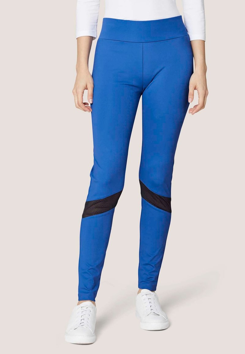 Blauwe leggings met een hoge tailleband, voorzien van zwarte meshpanelen bij de knieën en een gladde, rekbare stof. G gedragen met witte sneakers.