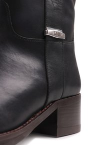 Κοντινή λήψη μαύρου δερμάτινου μποτάκι αστραγάλου με ξύλινο τακούνι, λεπτομερείς ραφές και μια μικρή ασημένια μεταλλική ετικέτα στο πλάι.