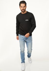 Black sweatshirt with a round neckline and ribbed cuffs, worn over light blue jeans. Features a small embroidered logo on the chest.
