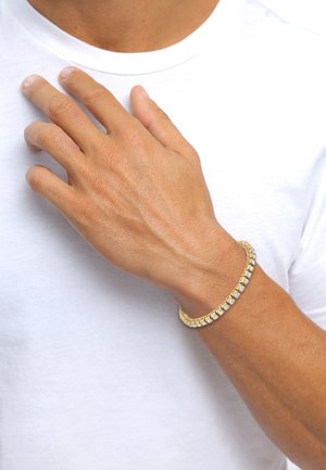 Goldfarbener Armband mit quadratischen Gliedern, die abwechselnd mit klaren Edelsteinen versehen sind, getragen an einer Hand vor einem schlichten weißen Hemd-Hintergrund.