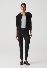 Gilet noir en peluche sur un haut gris à manches longues, associé à un pantalon noir à cordon de serrage avec poches zippées, et des chaussures noires à enfiler.