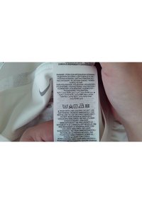 Étiquette de soin sur tissu blanc Nike montrant les instructions de lavage, le matériau polyester et les symboles de soin tenus par une main sur fond blanc.