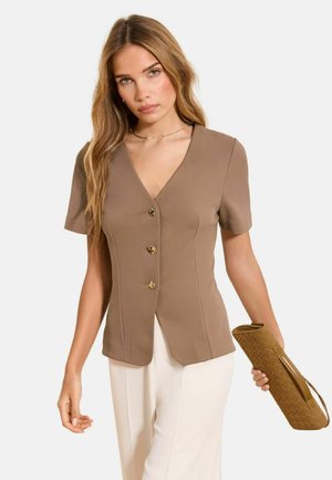 Femme portant un chemisier marron à manches courtes et boutonné, un pantalon blanc, tenant une pochette tissée beige sur un fond uni.