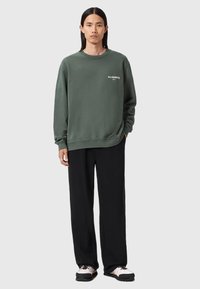 Zielony sweatshirt z ściągaczami przy rękawach i dole, z małym białym logo. Połączony z luźnymi czarnymi spodniami i casualowymi butami.