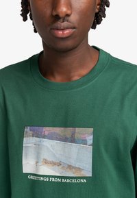 Grøn bomuldst-shirt med et rektangulært grafisk print af et landskab og teksten "GREETINGS FROM BARCELONA" i hvid nært bunden.