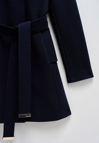 Manteau en laine bleu marine foncé avec manches longues, ceinture nouée à la taille et détail d'étiquette métallique à l'extrémité de la ceinture.