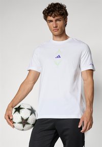T-shirt en coton blanc arborant un logo en forme de diamant violet et vert. Le mannequin tient un ballon de soccer à motifs, vêtu de shorts noirs.
