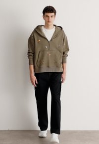 Sudadera verde oliva con bolsillo frontal, con bordado de animales, combinada con pantalones negros con cordones y zapatillas blancas.