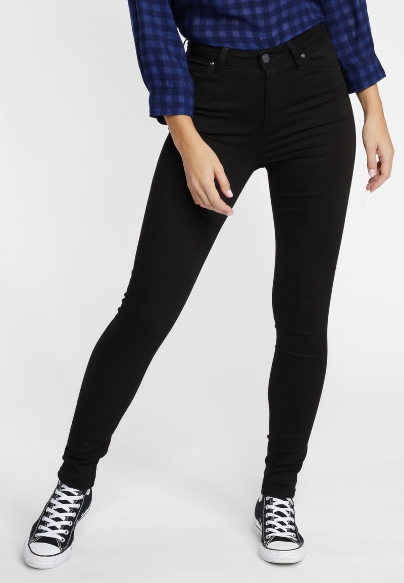 Personne portant un pantalon noir ajusté, une chemise bleue à carreaux et des baskets noir et blanc, debout les jambes écartées sur fond blanc.