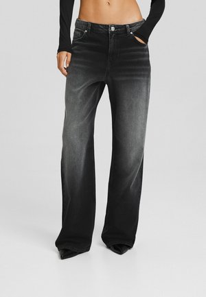 Weit geschnittene, ausgewaschene schwarze Jeans, getragen von einer stehenden Person mit einer Hand in der Tasche und freiem Bauch, vor einem einfachen grauen Hintergrund.