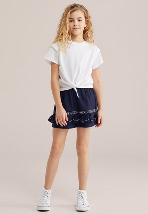 Dekle z dolgimi kodrastimi blond lasmi, oblečeno v belo majico, ki je zavezana v vozel, mornarsko modro krilo z volani in belimi šivi ter bele superge s težkim podplatom.