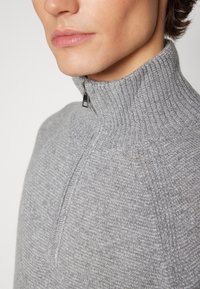 Pull en maille gris avec un col haut, doté d'une fermeture éclair à moitié zippée et d'une surface texturée. Gros plan sur le col et le détail de la fermeture éclair.