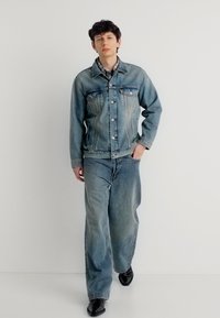 Νεαρός άνδρας που περπατά φορώντας ανοιχτό μπλε τζιν μπουφάν και τζιν παντελόνι με φαρδιά πόδια, με μαύρα μυτερά παπούτσια, σε απλό λευκό φόντο.