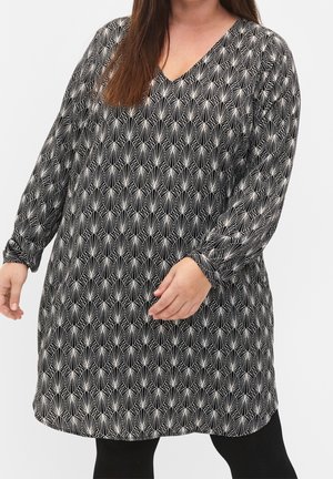 Kobieta ubrana w tunikę z długim rękawem o czarno-białym wzorze, z dekoltem w serek, w połączeniu z czarnymi legginsami, stoi na jednolitym tle.